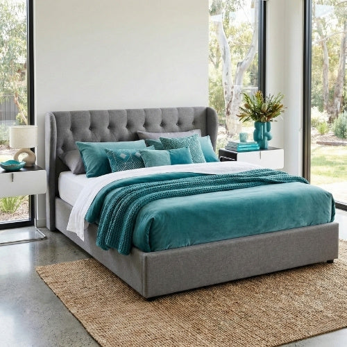Rivercity Furniture grey tufted bed frame in a modern Australian bedroom. Styled with teal bedding, pillows, and decor accents. Features large windows, polished concrete floors, and a jute rug.